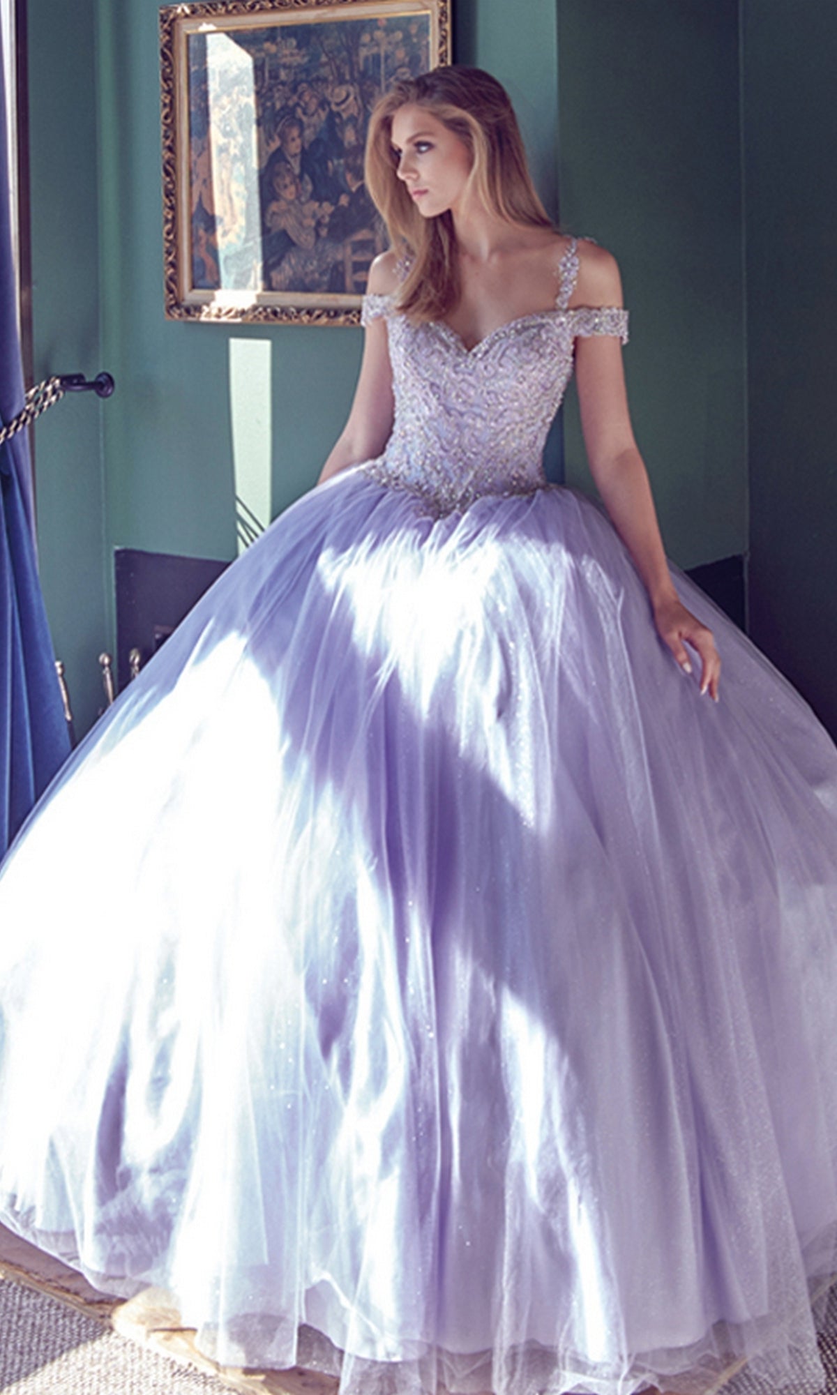 A woman stands in a sunlit room wearing the Juliet 1430 Cold-Shoulder Glitter Quinceanera Dress with a beaded bodice and off-the-shoulder straps.